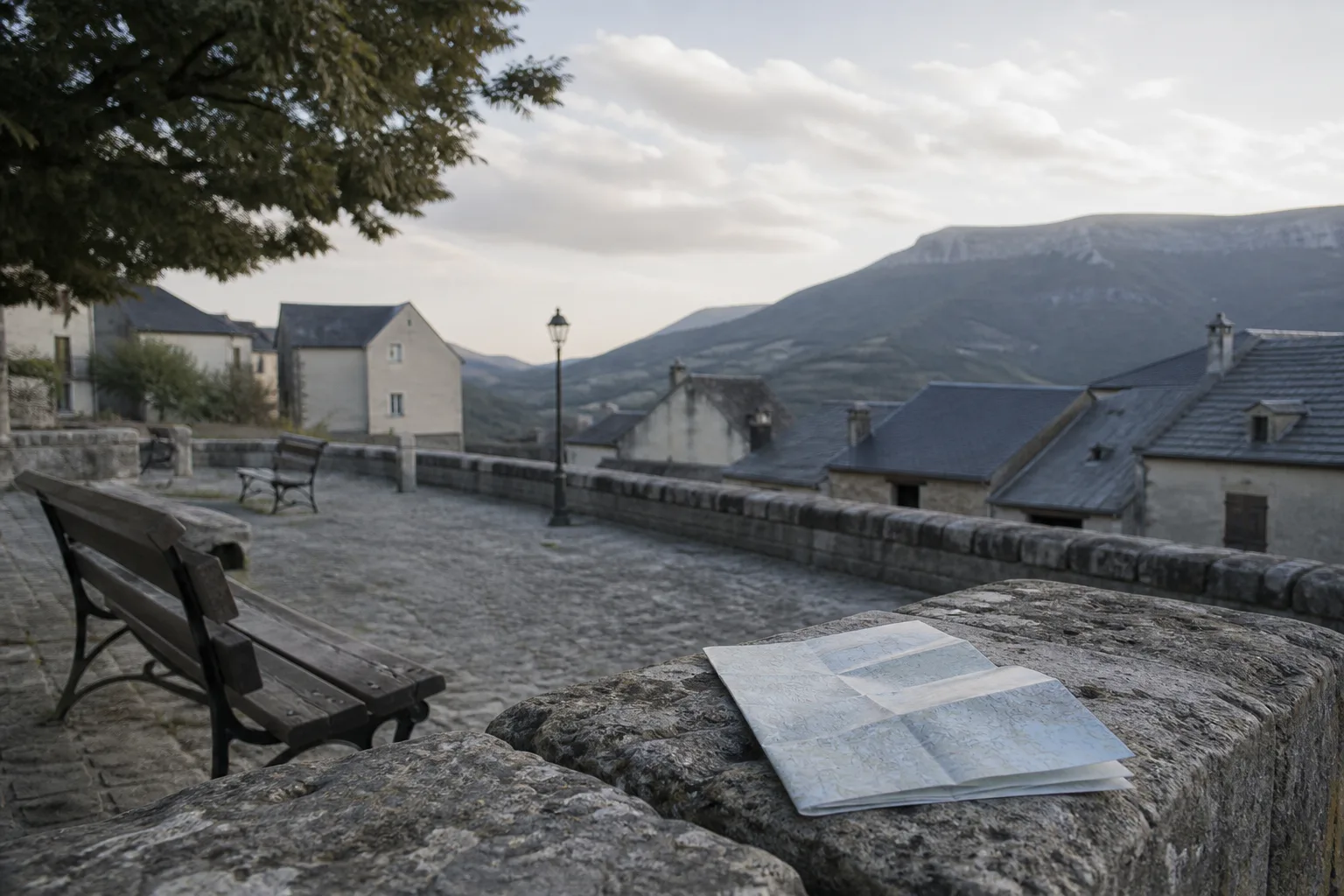 Town square in soft afternoon light with a distant ridge visible beyond rooftops, empty benches in the foreground, a folded map resting unused on a stone ledge, 1400x900