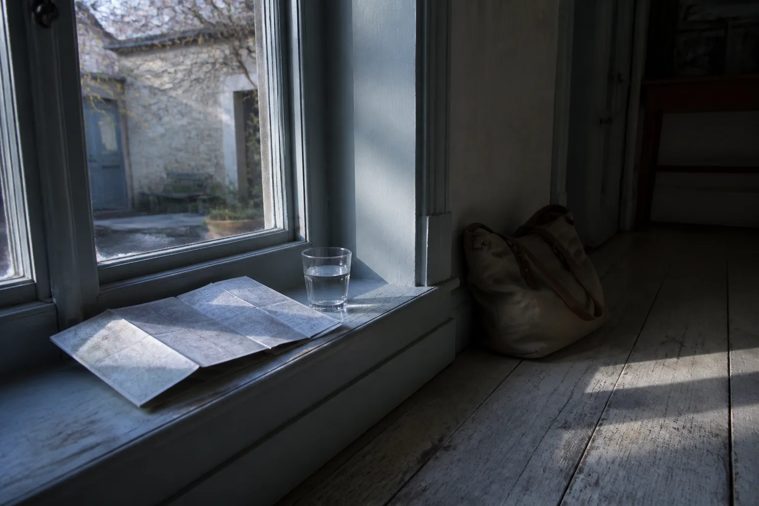 Folded paper map resting on a painted windowsill beside a glass of water, late afternoon light cutting across worn floorboards, a canvas day bag slumped against the wall, quiet courtyard visible through the glass, 1400x900